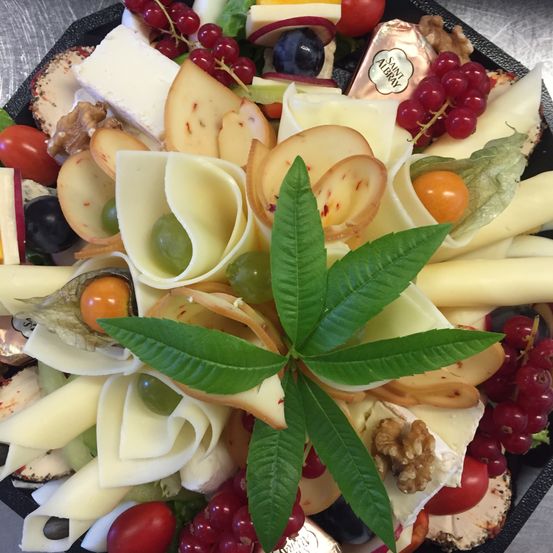 Ein Käseplatten-Arrangement zeigt verschiedene Käsesorten, einschließlich einer großen Blattform mit Trauben, umgeben von roten Beeren, Tomaten und Nüssen, alles auf einem schwarzen Tablett angeordnet.