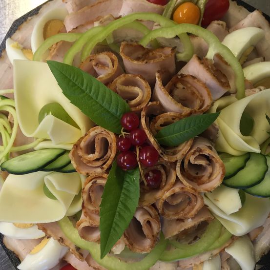 Ein Teller mit verschiedenen Fleisch- und Gemüsesorten, einschließlich geschnittenem Schinken, Käse, Gurken und Kirschtomaten, ist kunstvoll mit grünen Blättern arrangiert.
