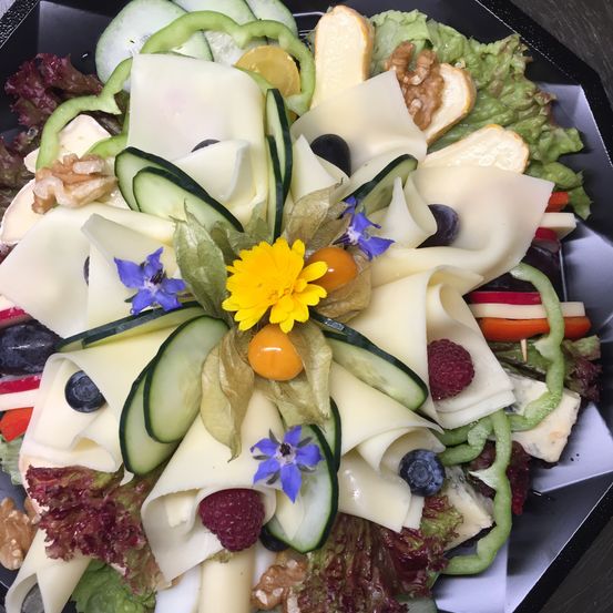 Eine runde Platte mit verschiedenen Käsescheiben, Gurken und Blumen. Der Käse hat verschiedene Formen und ist mit Obst und Gemüse belegt.
