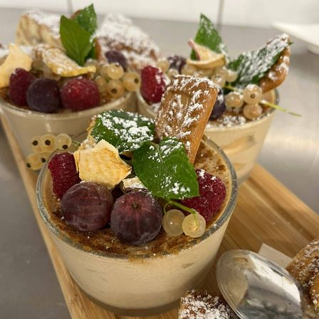 Vier Dessertbecher mit verschiedenen Toppings, einschließlich Beeren, grünen Blättern und Puderzucker, sind auf einem Holzbrett angeordnet. Jeder Becher hat eine andere Auswahl an Toppings.