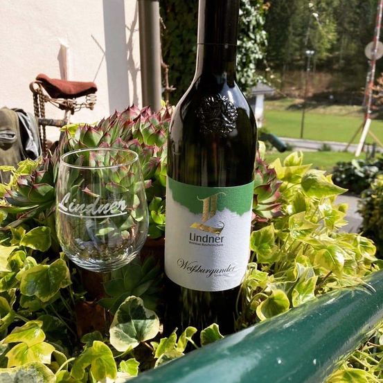 Eine Flasche Lindner-Wein steht zwischen grünen Blättern neben einem klaren Glas mit dem Wort 'Lindner'. Im Hintergrund befinden sich ein Fahrrad und eine Rasenfläche.