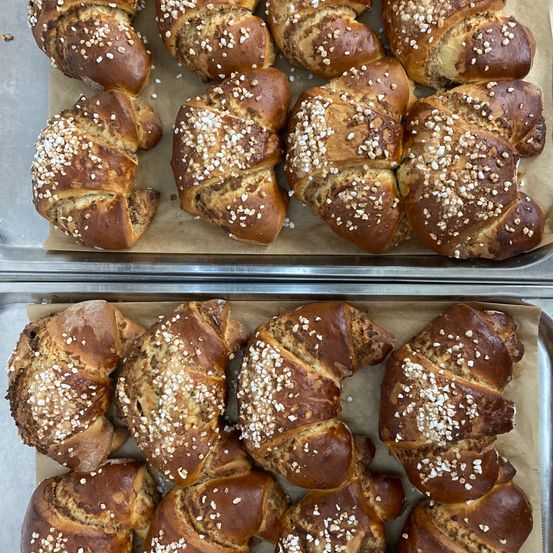 Zwei Tabletts mit frisch gebackenen, goldbraunen Brezeln, die mit Sesam bestreut sind, stehen auf einer weißen Oberfläche. Die Brezeln sind leicht gebogen und sehen außen knusprig aus.
