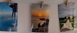 Drei Fotos hängen an einer weißen Wand. Das linke Foto zeigt eine Luftaufnahme einer Insel. Das mittlere Foto zeigt einen Sonnenuntergang über dem Ozean. Das rechte Foto zeigt ein Schloss und Gärten.