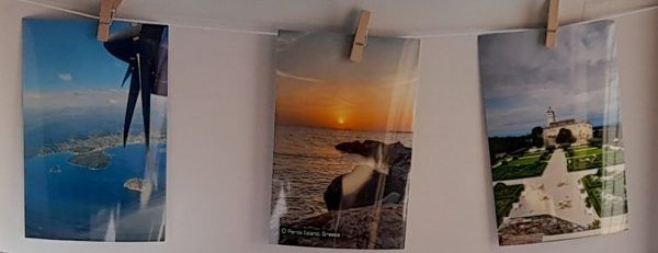 Drei Fotos hängen an einer weißen Wand. Das linke Foto zeigt eine Luftaufnahme einer Insel. Das mittlere Foto zeigt einen Sonnenuntergang über dem Ozean. Das rechte Foto zeigt ein Schloss und Gärten.