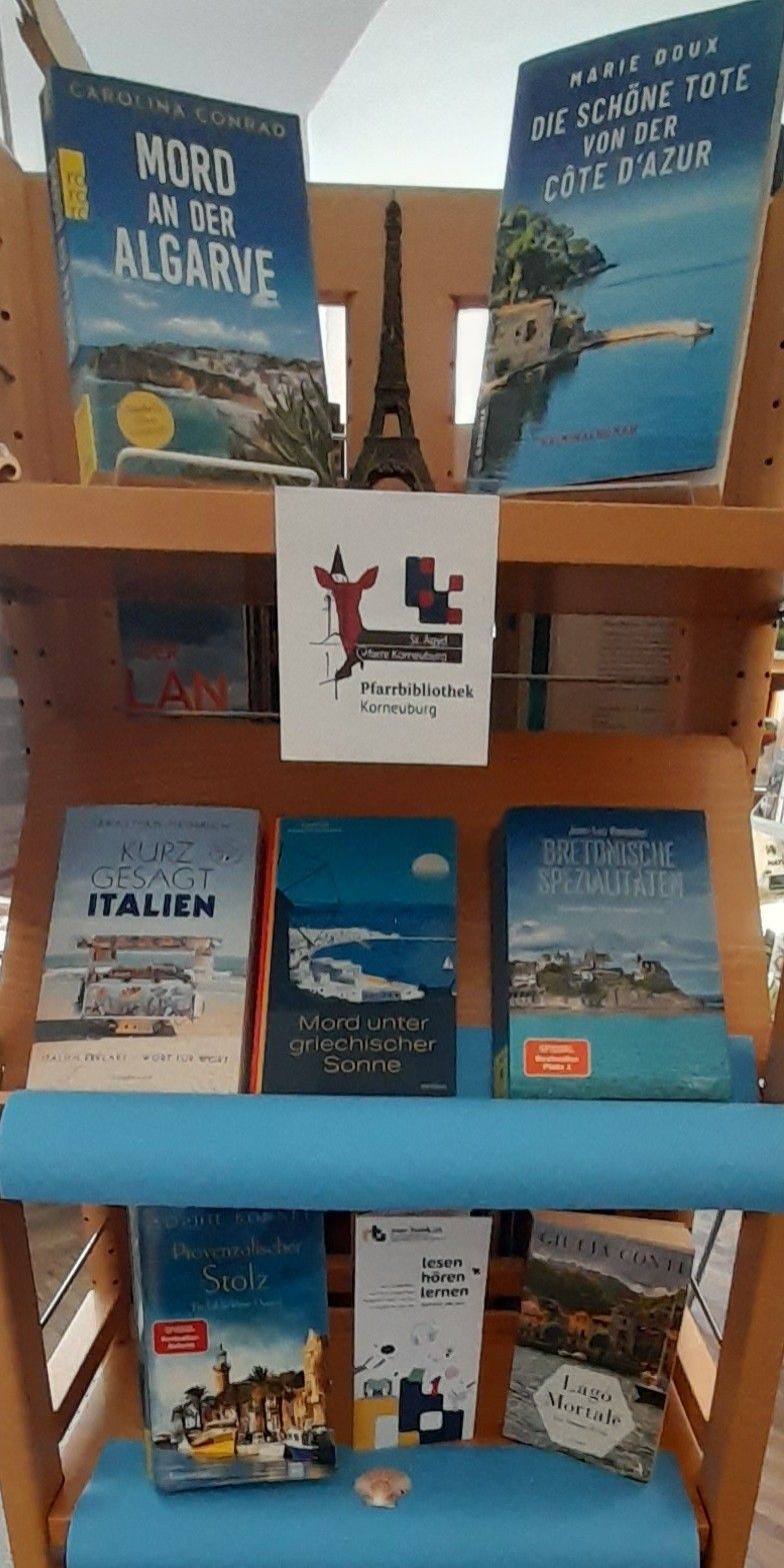 Ein Holzregal mit Büchern über Italien und griechische Landschaften. Drei Bücher sind ausgestellt, eines mit dem Titel 'Kurz Gesagt Italien', ein weiteres 'Mord unter griechischer Sonne' und das dritte 'Britanische Spezialitäten'.