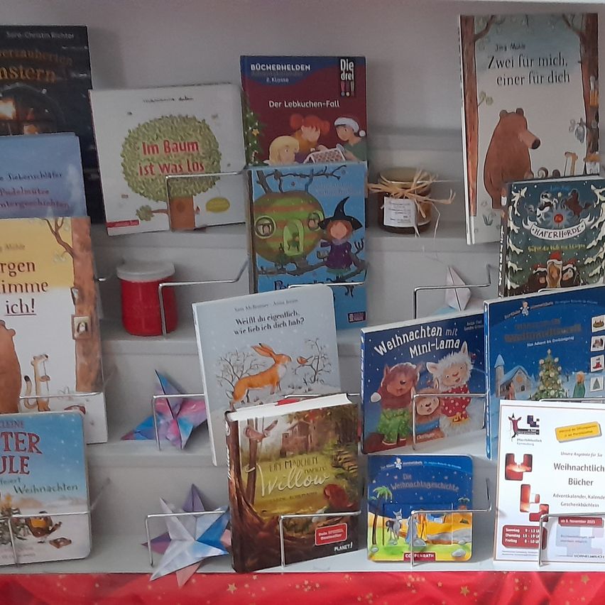 Ein weißer Regal zeigt mehrere Kinderbücher, einschließlich Titel wie 'In Baum ist was los' und 'Die drei drei.' Ein roter Behälter und dekoratives Origami sind ebenfalls vorhanden.