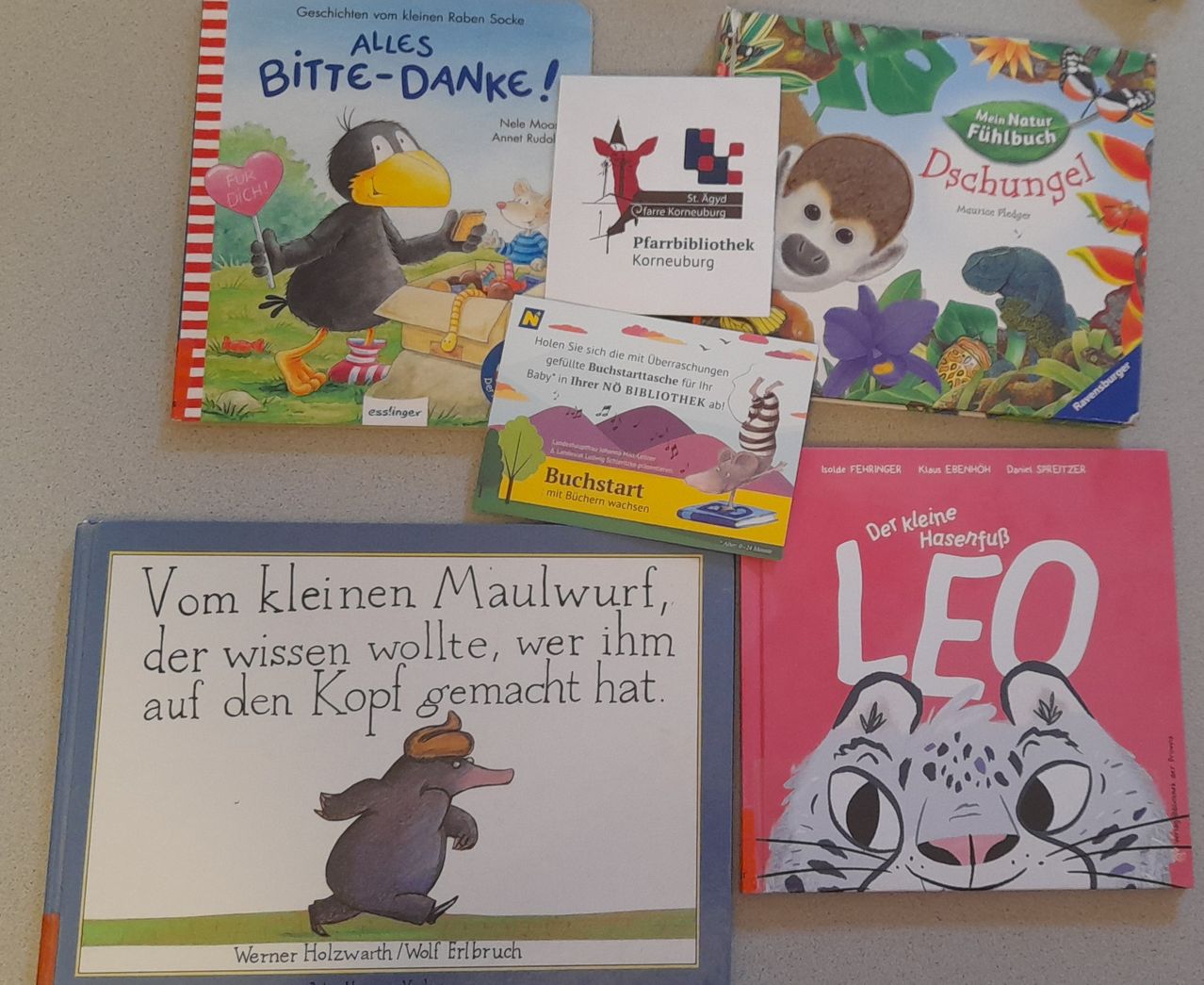 Mehrere Kinderbücher sind ausgestellt, darunter 'Alles Bitte-Danke!', 'Vom kleinen Maulwurf' und 'Det kleine Hasenfüß'. Die Bücher sind bunt mit Cartoon-Illustrationen. Eine Bibliothekskarte ist ebenfalls sichtbar.