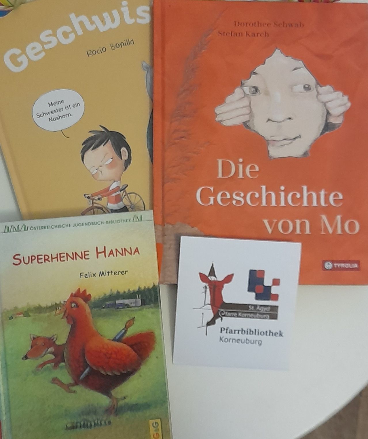 Vier Bücher sind ausgestellt. 'Gesamt' von Rocio Bonilla zeigt einen Jungen, der Fahrrad fährt. 'Die Geschichte von Mo' zeigt ein Kind, das sein Gesicht bedeckt. 'Superhennen Hanna' von Felix Mitterer zeigt einen Hahn und einen Fuchs. Eine kleine Karte mit 'Pfarrbibliothek Korneburg'.
