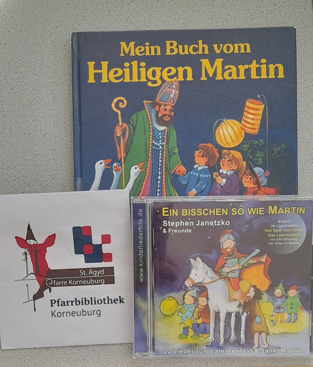 Zwei Bücher und ein CD-Case sind ausgestellt. Das Buch mit dem Titel 'Mein Buch vom Heiligen Martin' zeigt eine Illustration des Heiligen Martin. Darunter steht auf einem weißen Papier 'Pfarrbibliothek Korneburg'. Ein CD-Case mit 'Ein Bisschen so wie Martin' von Stephen Janetzko und Freunden zeigt einen Mann auf einem Pferd.