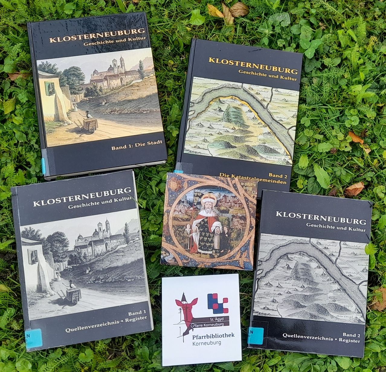 Fünf Bücher sind auf dem Gras angeordnet. Drei Bücher sind mit 'Klosterneuburg Geschichte und Kultur' betitelt. Zwei Bücher haben Karten und das Wort 'Band' auf dem Cover. Eine kleine Karte mit einem roten Design ist zwischen den Büchern platziert.