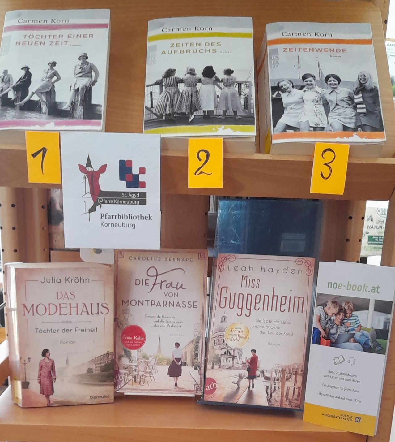 A wooden bookshelf displays several books, numbered 1 to 3. Book 1 is titled 'Tochter Einer Neuen Zeit'. Book 2 is titled 'Zeiten Des Aufbruchs'. Book 3 is titled 'Zeitenwende'. Below, books 'Das Modehaus', 'Die Frau Von Montparnasse', and 'Miss Guggenheim' are also displayed.