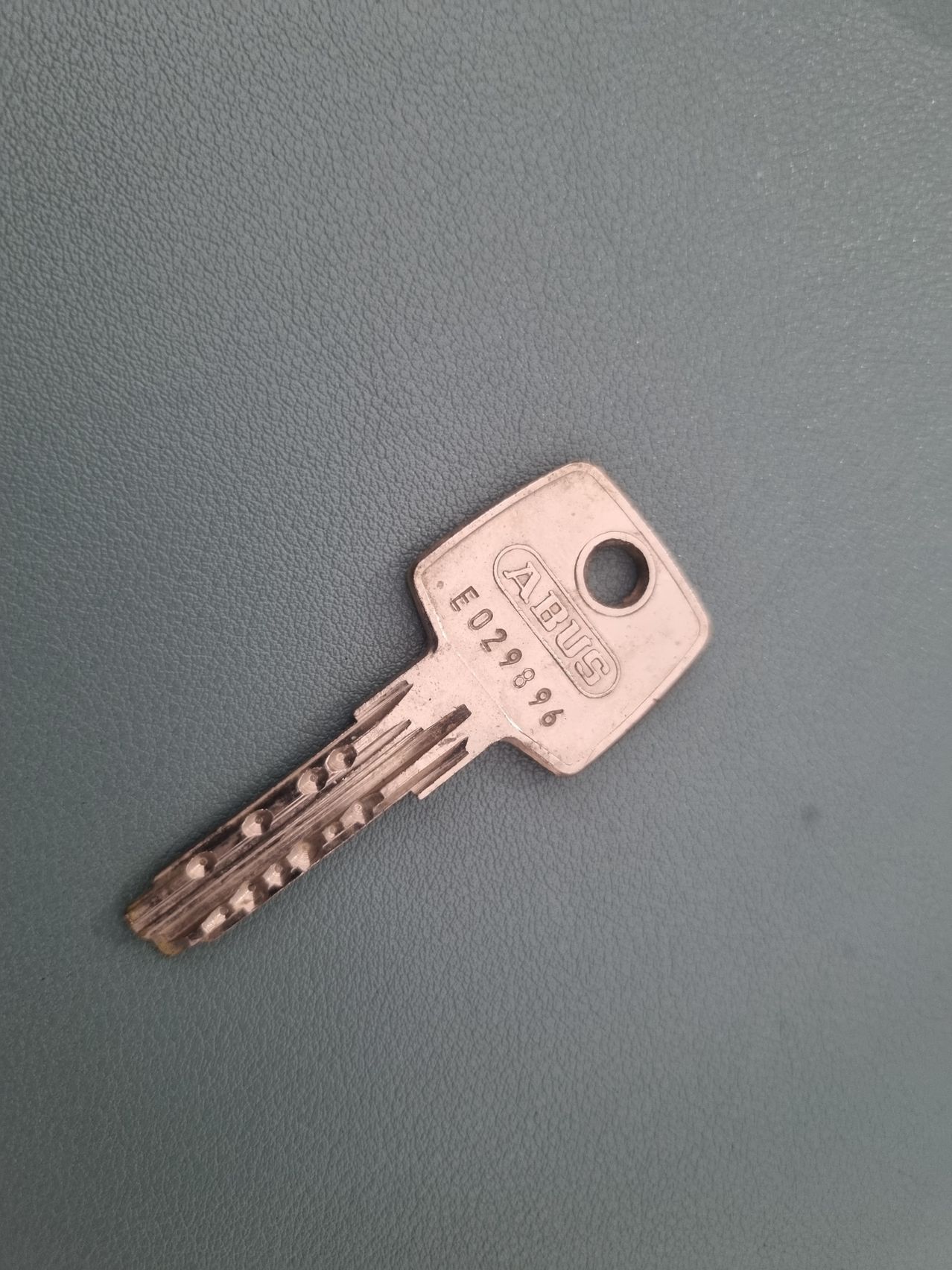 Auf einer grauen Lederoberfläche liegt ein alter silberner Schlüssel mit 'ABUS' und der Nummer 'E029896'. Auf ihm sind Abnutzungsspuren zu sehen.