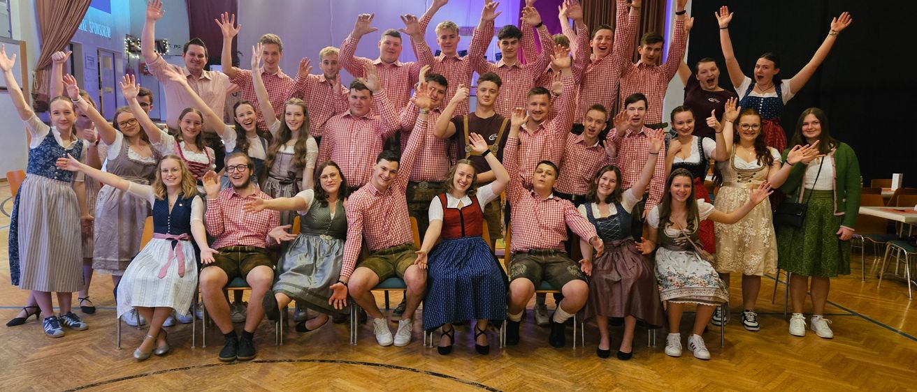 Eine Gruppe von Menschen in traditioneller Kleidung, einige stehend und andere sitzend, posiert für ein Foto mit erhobenen Händen in einem dekorierten Saal.