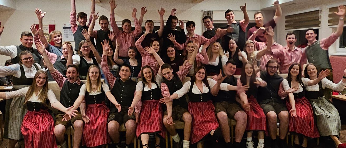 Eine Gruppe von Menschen in traditioneller Kleidung posiert für ein Foto mit erhobenen Händen, vermutlich bei einer gesellschaftlichen Veranstaltung. Der Raum hat Holzböden und Lampen an der Decke.