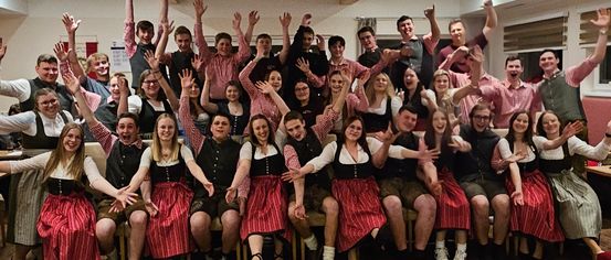 Eine Gruppe von Menschen in traditioneller Kleidung posiert für ein Foto mit erhobenen Händen, vermutlich bei einer gesellschaftlichen Veranstaltung. Der Raum hat Holzböden und Lampen an der Decke.