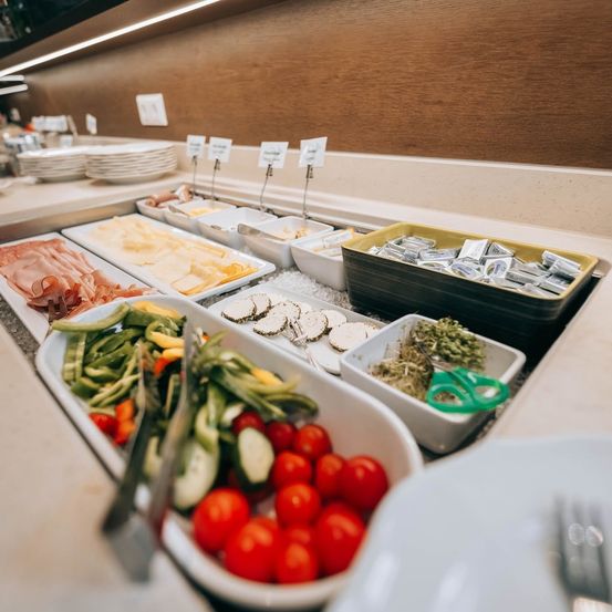 Ein Buffettisch mit verschiedenen Lebensmitteln wie geschnittenem Fleisch, Käse, Gemüse und Gewürzen, alles ordentlich in weißen Behältern angeordnet.