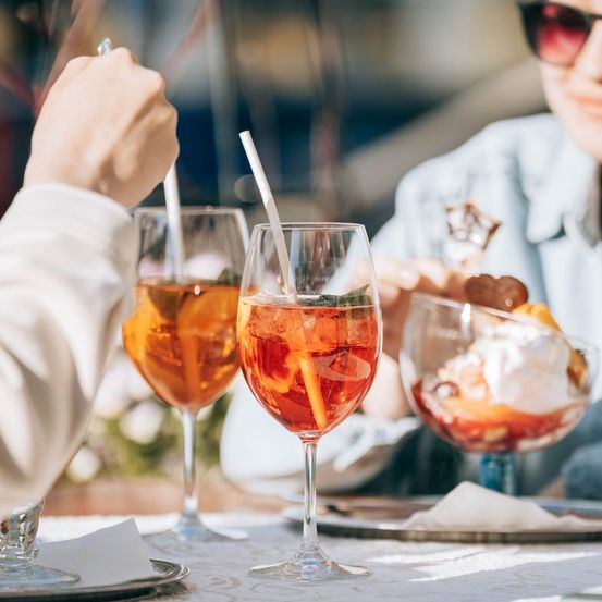 Zwei Personen sitzen an einem Tisch, eine hält ein Glas mit einem roten Getränk und einem Strohhalm, die andere mit einem Dessert in einem Glasbecher.