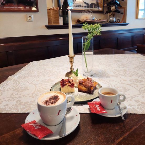 Ein Tisch mit weißer Tischdecke, auf dem sich eine Platte mit Desserts, zwei Tassen Kaffee, eine Kerze und eine Vase mit Blumen befindet.