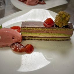 Ein Stück geschichteter Kuchen mit Schokolade, Pistazie und grüner Füllung, garniert mit Kirsche und kirschähnlicher Frucht, begleitet von einer Kugel rosa Eiscreme und Kirschscheiben auf einem weißen Teller.