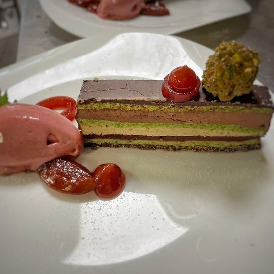 Ein Stück geschichteter Kuchen mit Schokolade, Pistazie und grüner Füllung, garniert mit Kirsche und kirschähnlicher Frucht, begleitet von einer Kugel rosa Eiscreme und Kirschscheiben auf einem weißen Teller.