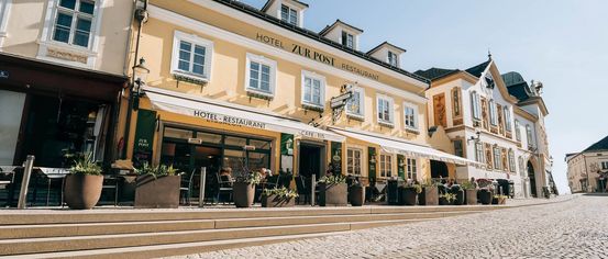 A yellow building with the name Hotel Zur Post Restaurant, featuring a white awning and several potted plants in front. There are people sitting outside at tables.
