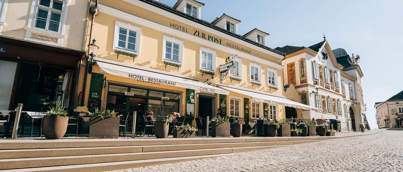 Ein gelbes Gebäude mit dem Namen Hotel Zur Post Restaurant, mit einer weißen Markise und mehreren Topfpflanzen davor. Es gibt Leute, die draußen an Tischen sitzen.