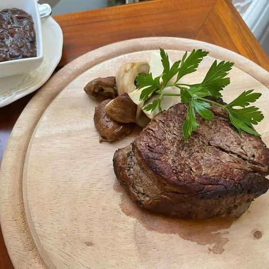 Ein Holzteller mit einem Steak, das mit Pilzen und Petersilie garniert ist, begleitet von einer Schüssel Sauce.