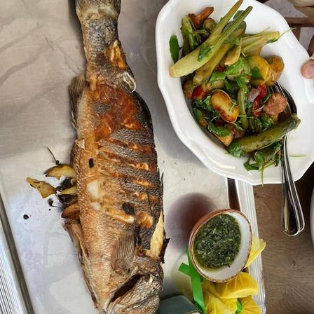 Ein ganzer gegrillter Fisch liegt auf einem Metalltablett mit einer Beilage aus Gemüse und einer kleinen Schüssel grüner Sauce.