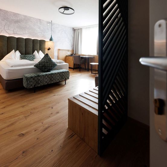 Ein modernes Schlafzimmer mit Holzboden, einem großen Bett und einer Bank. Ein Schreibtisch und ein Stuhl stehen in der Ecke, und ein Fenster mit Vorhängen befindet sich hinter dem Schreibtisch.