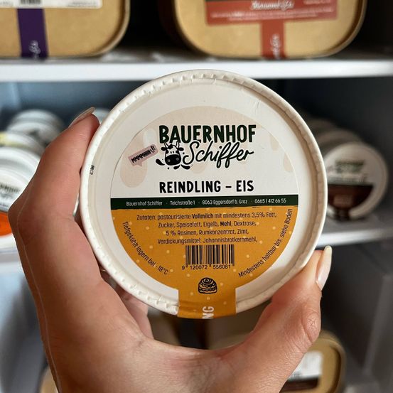 Eine Hand hält einen weißen und gelben Behälter mit der Aufschrift 'Bauernhof Schiffer Reindling - Eis' und einem Kuhlogo. Der Behälter ist mit einer cremigen Substanz gefüllt und hat einen Barcode und Nährwertinformationen.