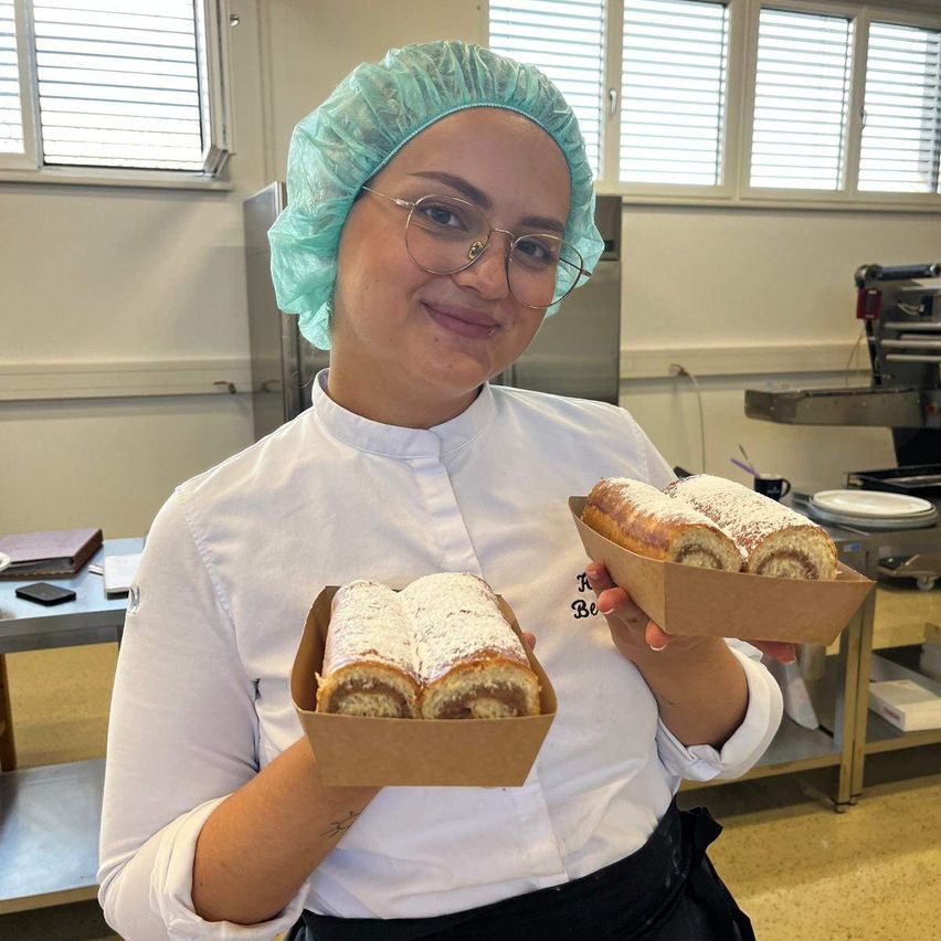 A woman in a chef's uniform holds two boxes of pastries, smiling for a photo in a kitchen.