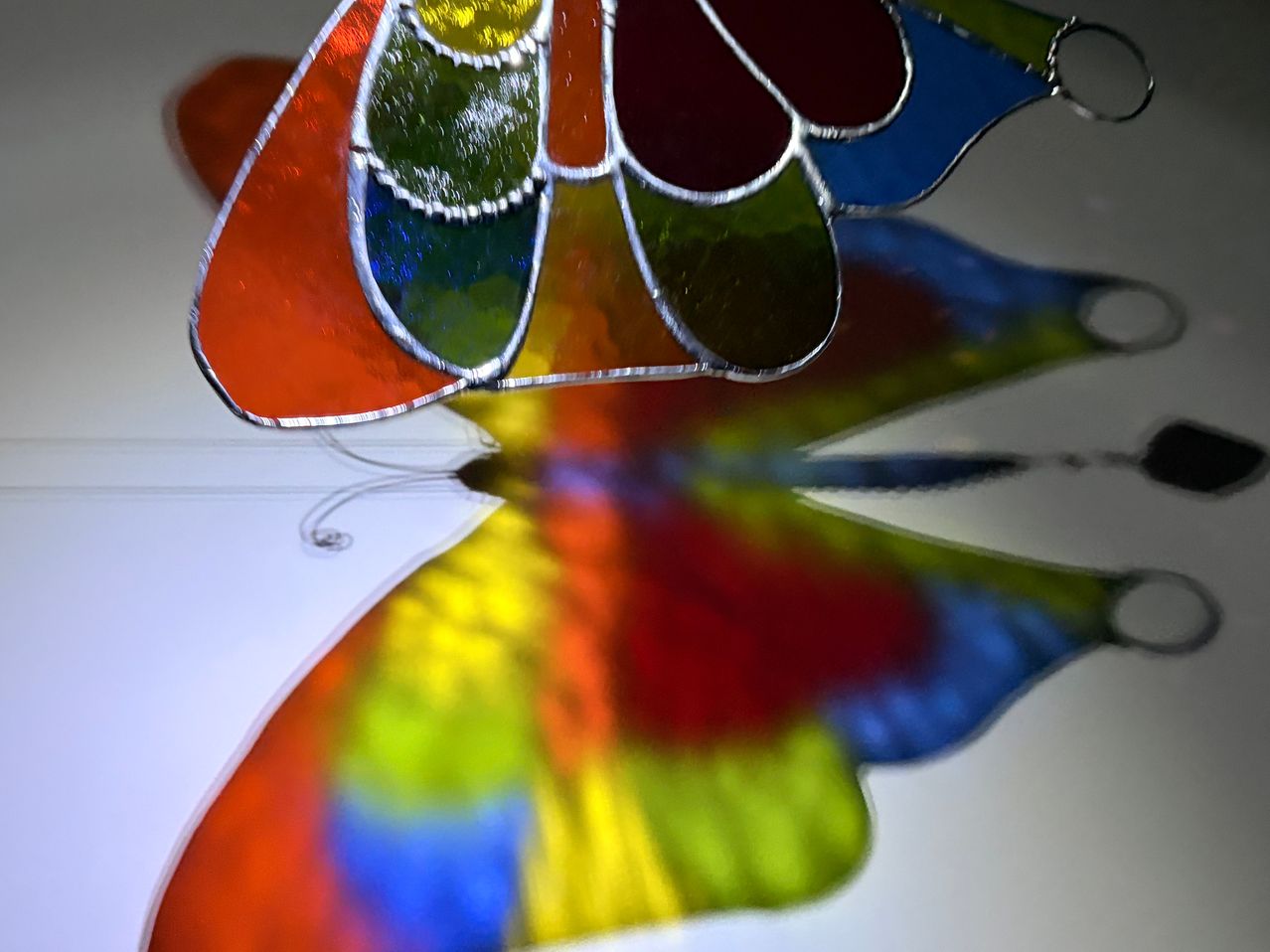 Ein farbenfrohes Buntglas-Schmetterling mit leuchtenden Rot-, Blau- und Grüntönen, reflektiert auf einer weißen Oberfläche.