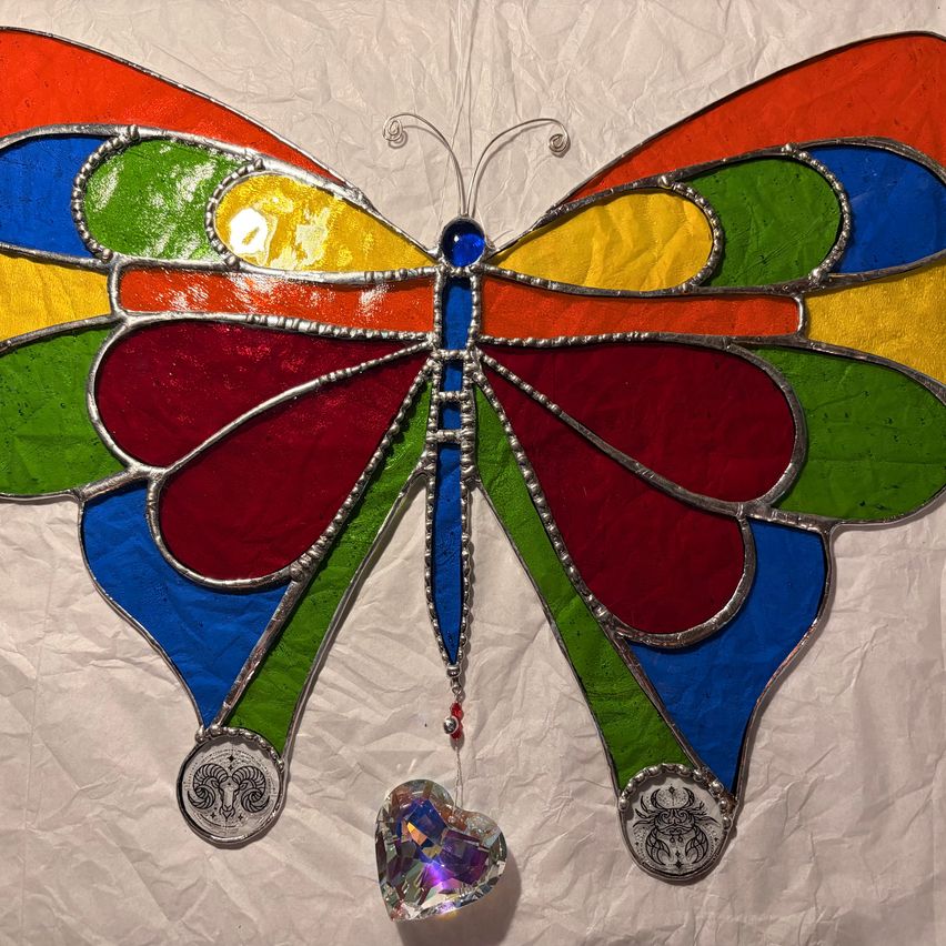 Ein farbenfroher Glaskunst-Schmetterling mit mehrfarbigen Flügeln und einer hängenden Herz-Anhänger. Die Flügel sind mit silbernen Akzenten und Symbolen verziert, darunter ein Widder und ein Skorpion.