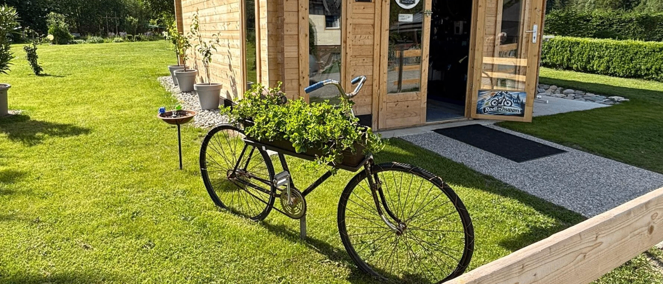 Ein altes Fahrrad mit einem Blumenkorb steht vor einem kleinen Holzladen mit Glastür und Markisen. Topfpflanzen befinden sich an der Seite.