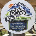 Ein rundes Schild mit dem Text Radlschuppen und einem Fahrradlogo. Die Öffnungszeiten sind Montag bis Freitag von 8:00-12:00, 14:00-17:00.