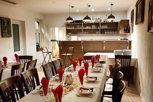 Der Essbereich eines Restaurants zeigt einen langen Tisch, der für eine Mahlzeit mit Tellern, Weingläsern und Servietten vorbereitet ist. Der Raum hat eine Holzbar, Regale mit Flaschen und gerahmte Bilder an den Wänden.