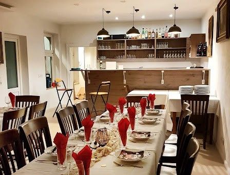 Der Essbereich eines Restaurants zeigt einen langen Tisch, der für eine Mahlzeit mit Tellern, Weingläsern und Servietten vorbereitet ist. Der Raum hat eine Holzbar, Regale mit Flaschen und gerahmte Bilder an den Wänden.