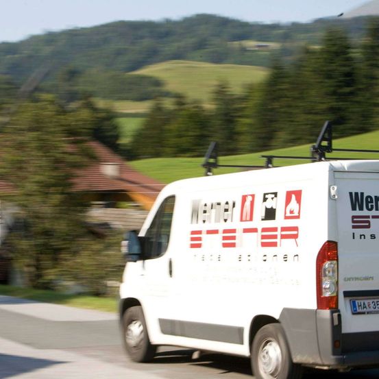 Ein weißer Lieferwagen mit Logo und Text auf der Seite fährt eine Straße hinunter, wobei der Hintergrund von Häusern und Bäumen verschwommen ist.