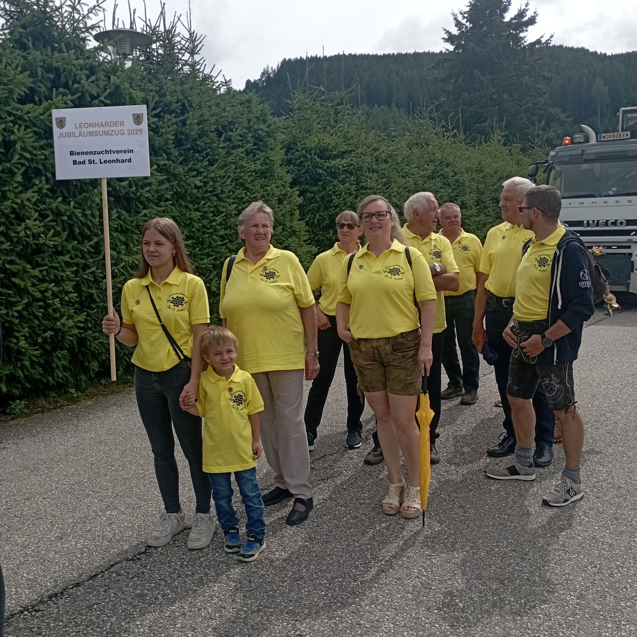 Eine Gruppe von Menschen in gelben Shirts und Shorts steht auf einer Straße. Eine Person in der Front hält ein Schild mit Text. Ein Lastwagen ist hinter ihnen geparkt.