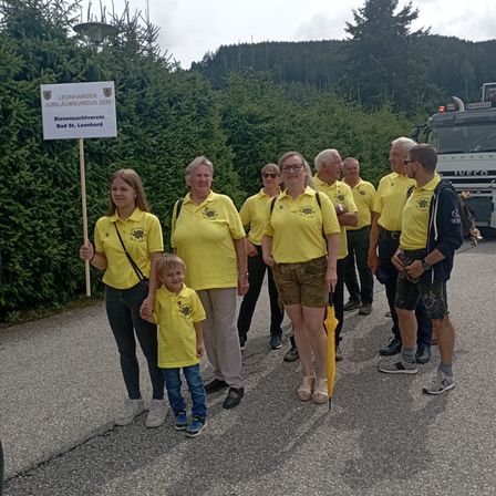 Eine Gruppe von Menschen in gelben Shirts posiert für ein Foto auf einer Straße. Ein Schild für Leonharder Jubiläumszug 2025 wird von einer Frau gehalten.