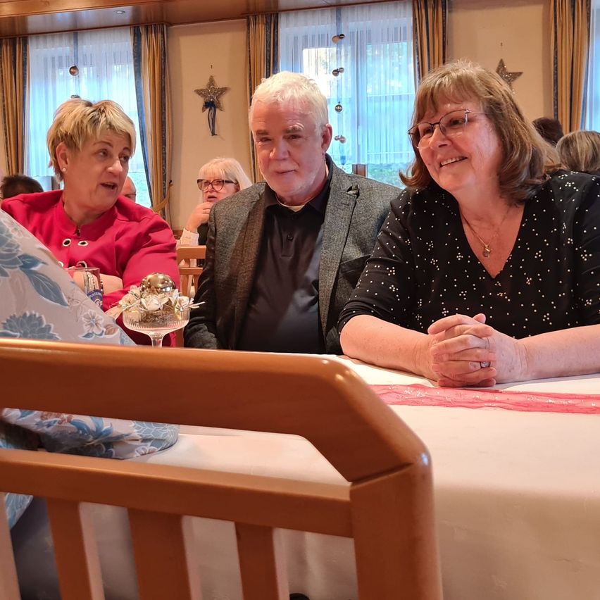 Drei Menschen sitzen an einem Tisch und lächeln für ein Foto. Eine Frau in Rot, ein Mann in Grau und eine weitere Frau in Schwarz. Der Tisch hat eine blumige Tischdecke.