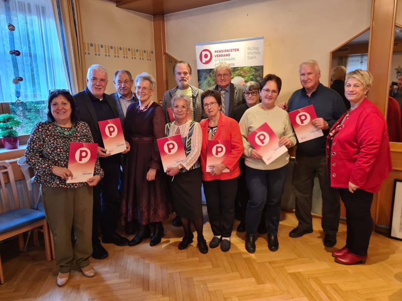 Eine Gruppe von Menschen, möglicherweise Rentner, stehen zusammen, lächeln und posieren für ein Foto. Sie halten rote Plakate mit dem Buchstaben P in der Hand. Hinter ihnen befindet sich ein Banner mit einem Logo und etwas Text.