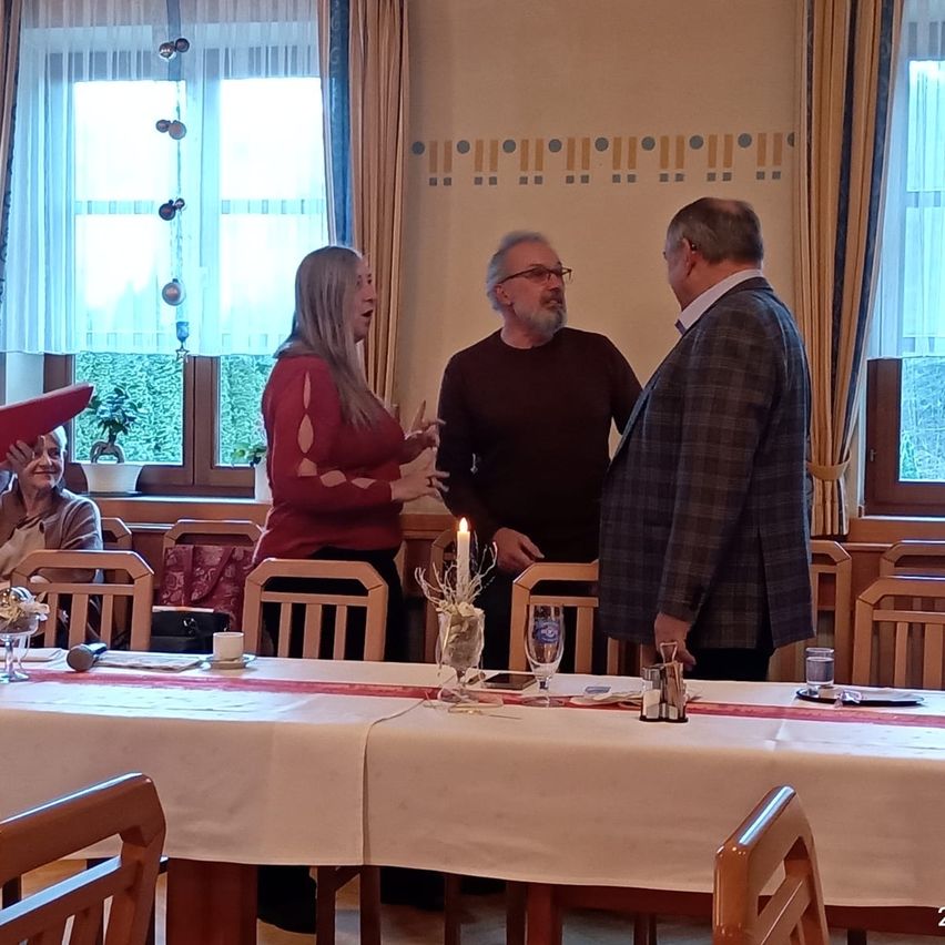 Eine Gruppe von Menschen versammelt sich um einen Tisch, wobei zwei Männer und eine Frau stehend plaudern. Der Tisch ist mit Kerzen, Gläsern und Tellern gedeckt. Eine Frau sitzt in einem Stuhl in der Nähe. Vor dem Fenster stehen Topfpflanzen.