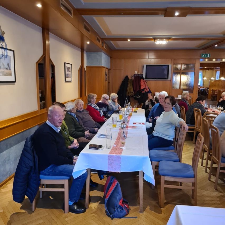 Eine Gruppe von Menschen sitzt an einem langen Tisch in einem Restaurant. Einige tragen Brillen, und es gibt Gläser und Teller auf dem Tisch. Holzstühle stehen um den Tisch herum. Ein Fernseher ist an der Wand montiert, und Bilderrahmen hängen an der Wand.