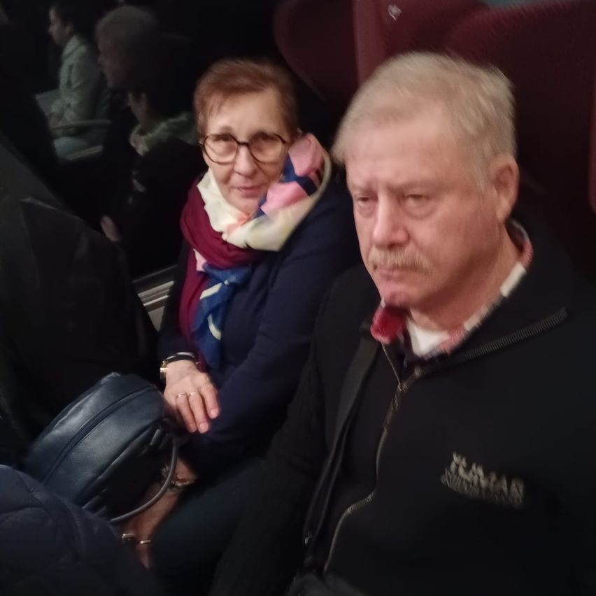 Ein älteres Paar sitzt in einem Fahrzeug, wahrscheinlich einem Zug. Die Frau trägt eine Brille und einen Schal, während der Mann einen Schnurrbart und eine schwarze Jacke hat.