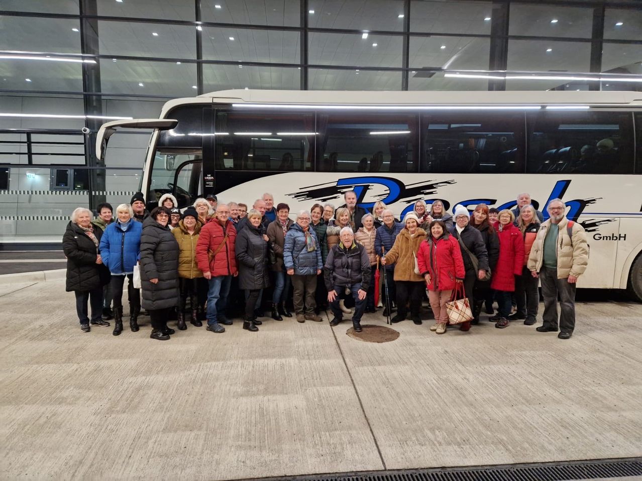 Eine Gruppe von Menschen steht vor einem weißen Bus in einem modernen Gebäude mit Glaswänden. Sie tragen Winterkleidung, und einige halten Taschen.