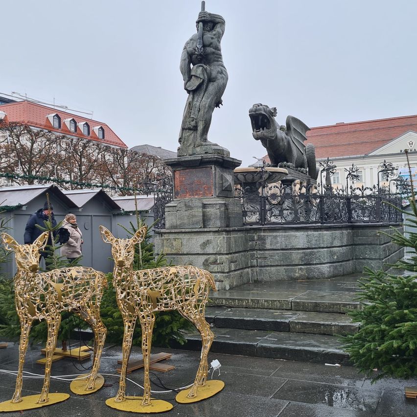 Eine Statue eines Mannes mit einem Schwert steht auf einem Podest. Dahinter spuckt eine Drachenstatue Feuer. Zwei goldene Hirschstatuen stehen davor, mit zwei Personen dahinter.