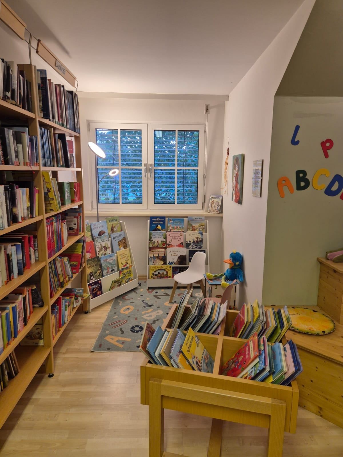 Eine gemütliche Kinderbücherei voller Bücher, eine Leseecke mit Stuhl und Lampe und Buchstabenwandkunst. Regale mit Büchern und ein Teppich mit Buchstaben.