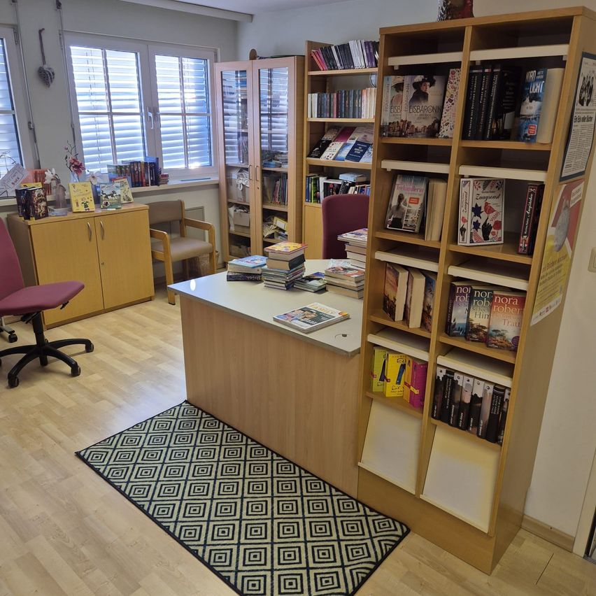 Ein Büroraum verfügt über einen Schreibtisch mit Büchern, einen gemusterten Teppich und hölzerne Bücherregale gefüllt mit Büchern. Ein Stuhl und ein Schrank befinden sich in der Nähe eines Fensters mit Jalousien.