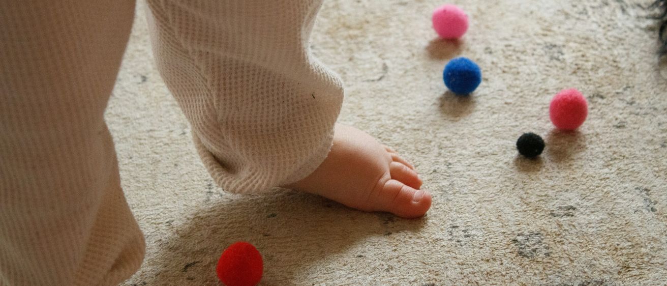 Ein Kinderfuß steht auf einem Teppichboden mit verschiedenen farbigen Filzbällen, darunter rot, pink, blau und schwarz.